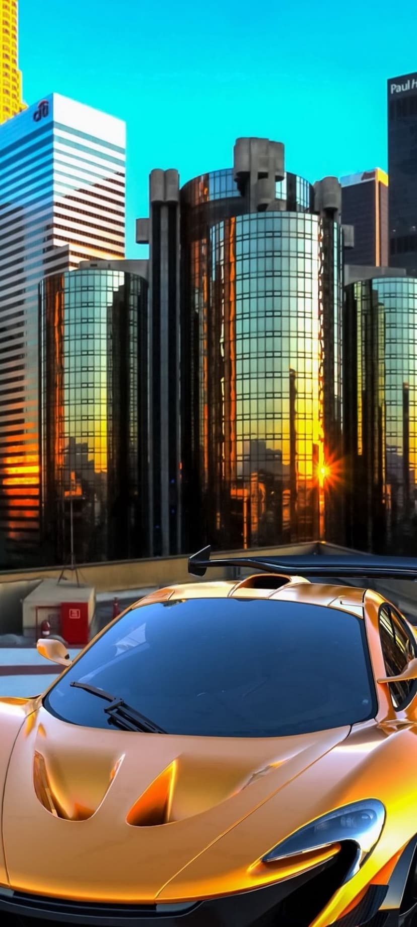 Golden McLaren P1 sports car parked on an urban street with sunset reflections on nearby glass skyscrapers.