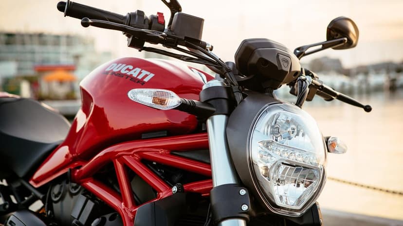 Red Ducati Monster motorcycle fuel tank and headlight close-up with a blurred marina background.