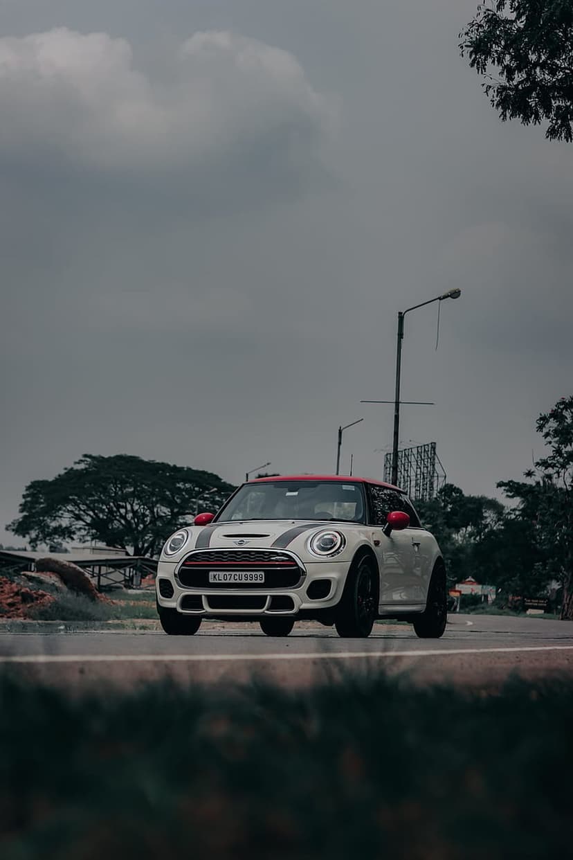 White Mini Cooper S with a red roof and racing stripes travels on a wet road under heavy, dark storm clouds.
