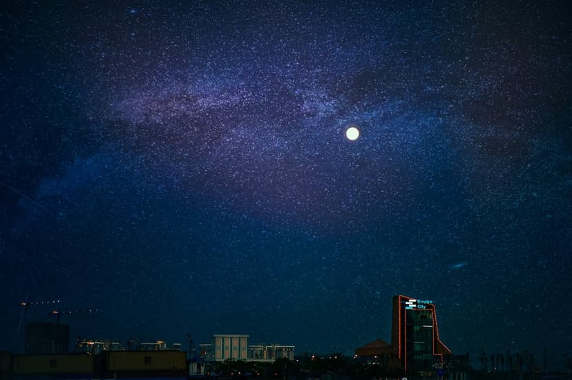 Illuminated skyscrapers stand beneath the swirling Milky Way galaxy and a bright moon in a deep purple night sky.
