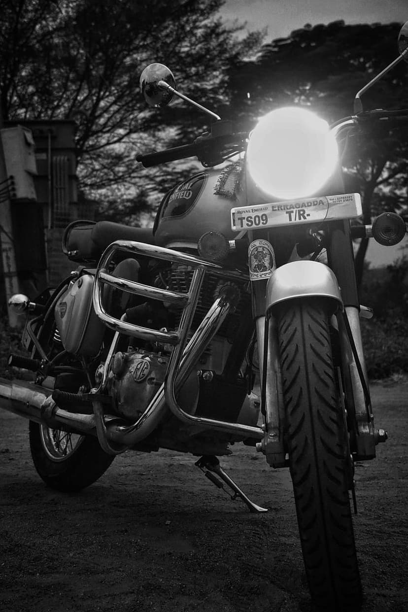 Royal Enfield motorcycle with a glowing headlight and chrome engine parts in a monochrome outdoor setting.