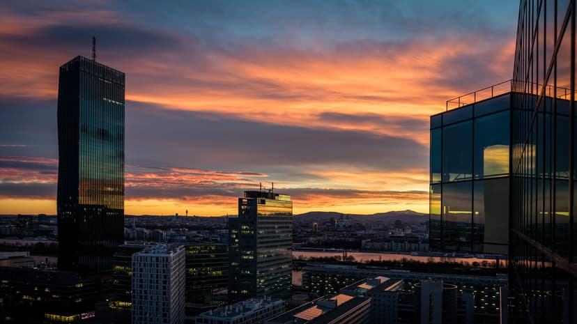 Glass skyscrapers reflect orange sunset clouds beside a river with distant mountains in this urban scene.
