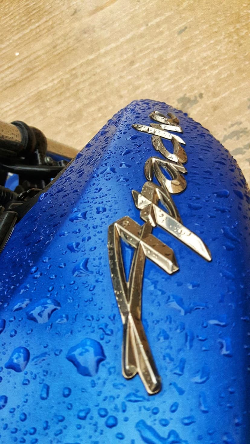 Blue motorcycle fuel tank with a metallic RX emblem and scattered water droplets against a wooden background.