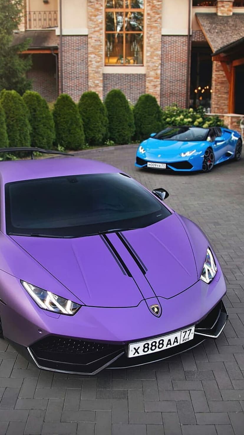 Purple and blue Lamborghinis parked on a stone driveway in front of a grand mansion with manicured hedges.