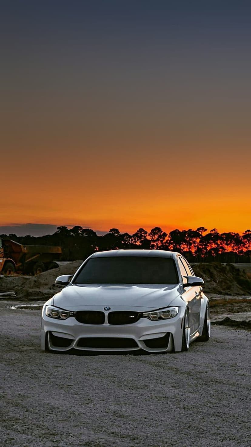 White BMW M4 parked on gravel with heavy construction machinery under a glowing orange and yellow sunset sky.