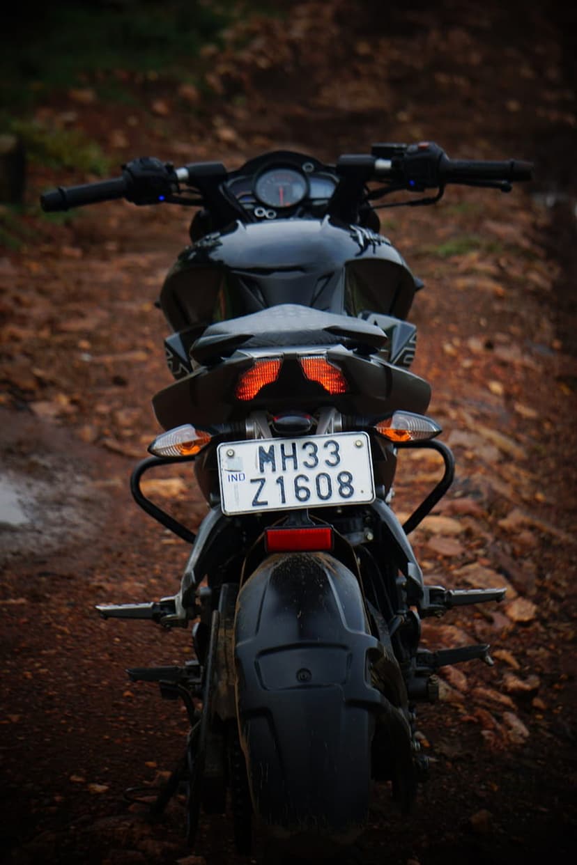 Black motorcycle rear fender with orange lights and a license plate on a gravel road with a blurred backdrop.