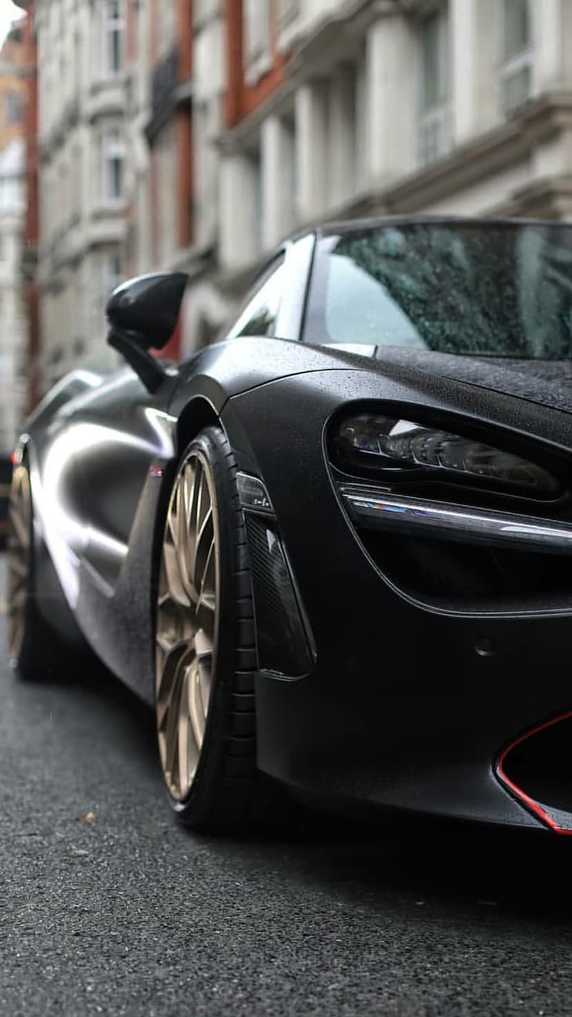 Black McLaren 720S supercar with gold wheels on a wet city street, featuring raindrops on glossy bodywork.