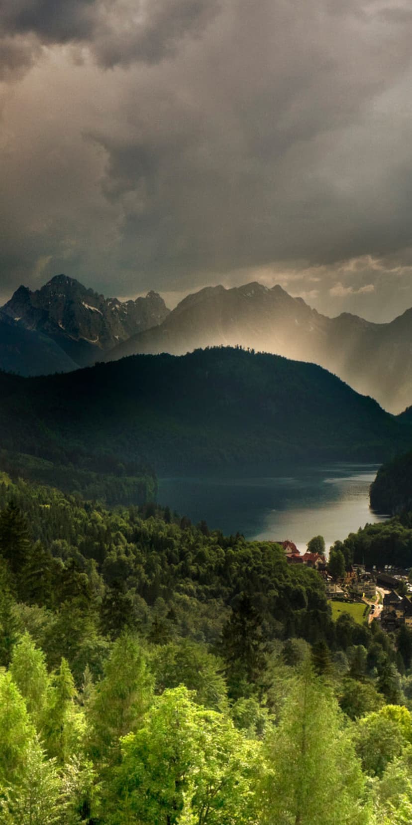 Emerald pine forests encircle a still lake and small village beneath towering, snow-dusted alpine mountain peaks.