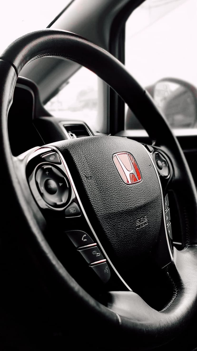 Honda steering wheel featuring a red central emblem, black leather texture, and silver multifunction controls.