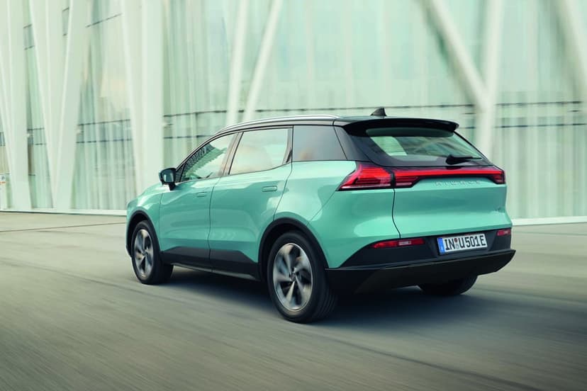 Teal Aiways U5 electric SUV driving past a modern white building with motion blur on the wheels and road.
