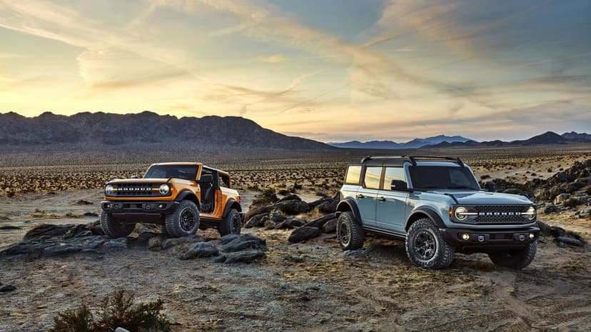 Two Ford Bronco SUVs on rocky desert terrain under a golden sunset sky with distant mountains and wispy clouds.