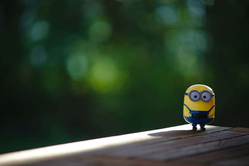Minion toy figurine stands on a wooden plank with a soft green blurred background and warm cinematic lighting.
