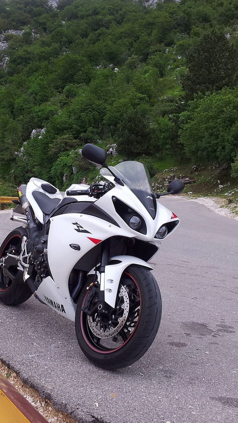 White Yamaha R1 sportbike parked on a paved mountain road curve surrounded by green trees and rocky cliffs.