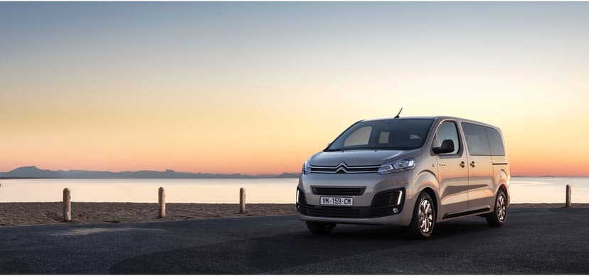 Silver Citroen van on a coastal road at sunset with calm ocean, sandy beach, and mountain silhouettes.