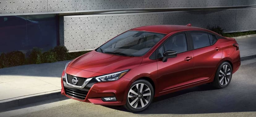 Red Nissan Versa sedan parked near a perforated metal building on a sunny day with sharp shadows.