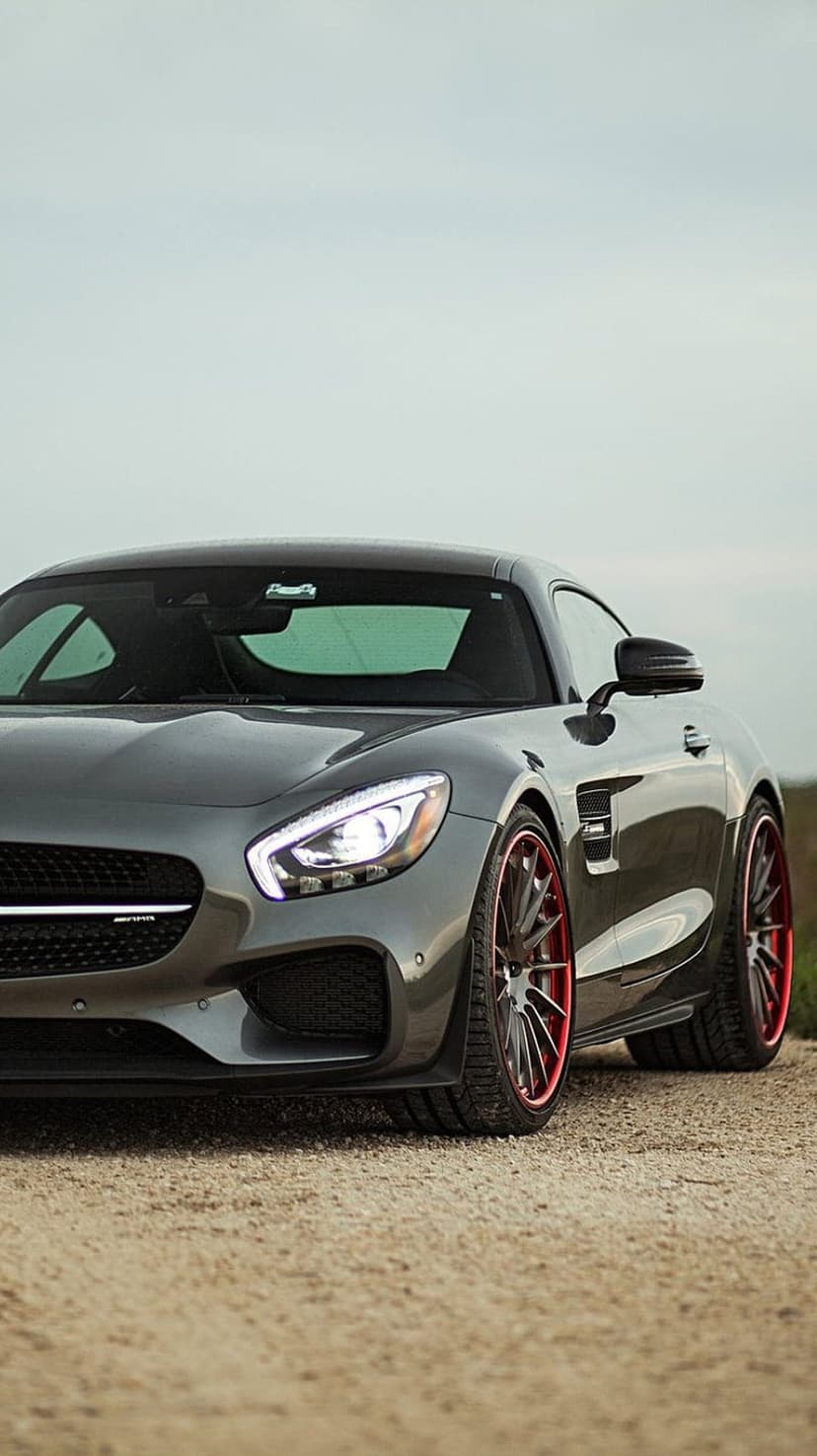 Mercedes-AMG GT S sports car with red-rimmed wheels and glowing headlights parked on a light gray gravel road.