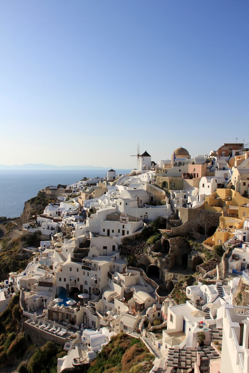 Whitewashed stone buildings with blue domes terrace down a steep volcanic cliff overlooking the Aegean Sea.