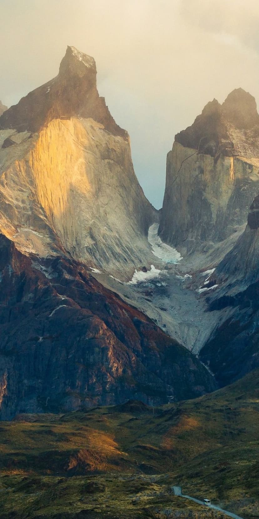 Jagged granite peaks of Torres del Paine tower over a lush green valley and winding road under warm sunlight.