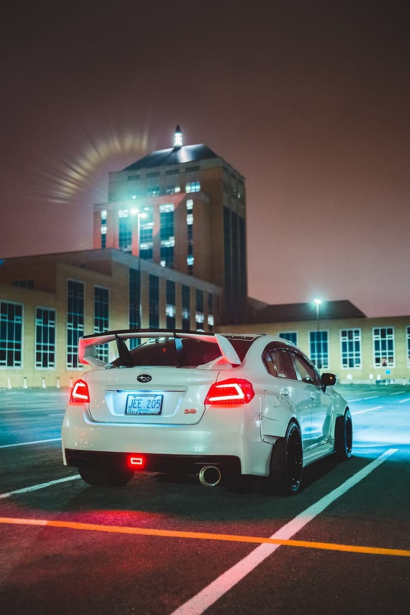 White Subaru WRX with widebody fenders and a large wing parked in a night lot with red taillights glowing.