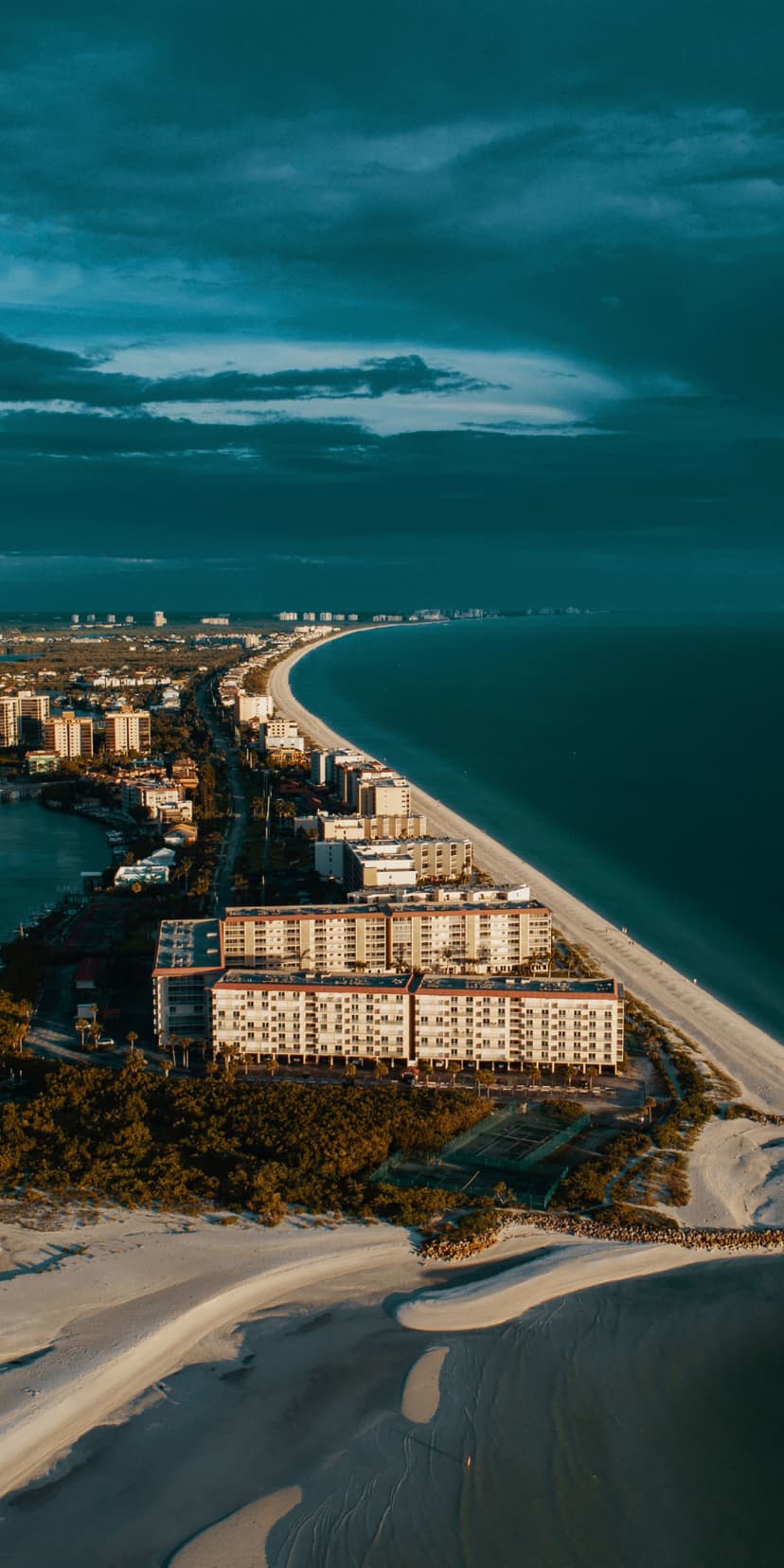 White sand beach curves along deep blue ocean water next to coastal hotels under a dramatic, cloudy sky.