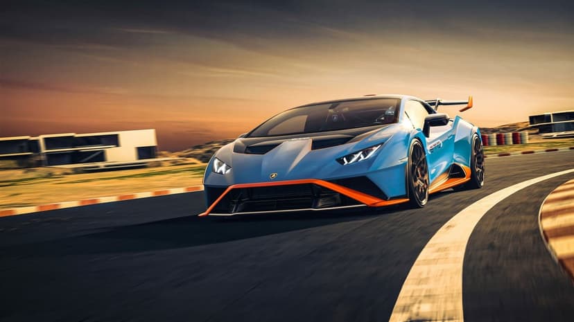 Blue Lamborghini Huracán STO with orange trim driving on a racetrack at sunset with motion-blurred background.