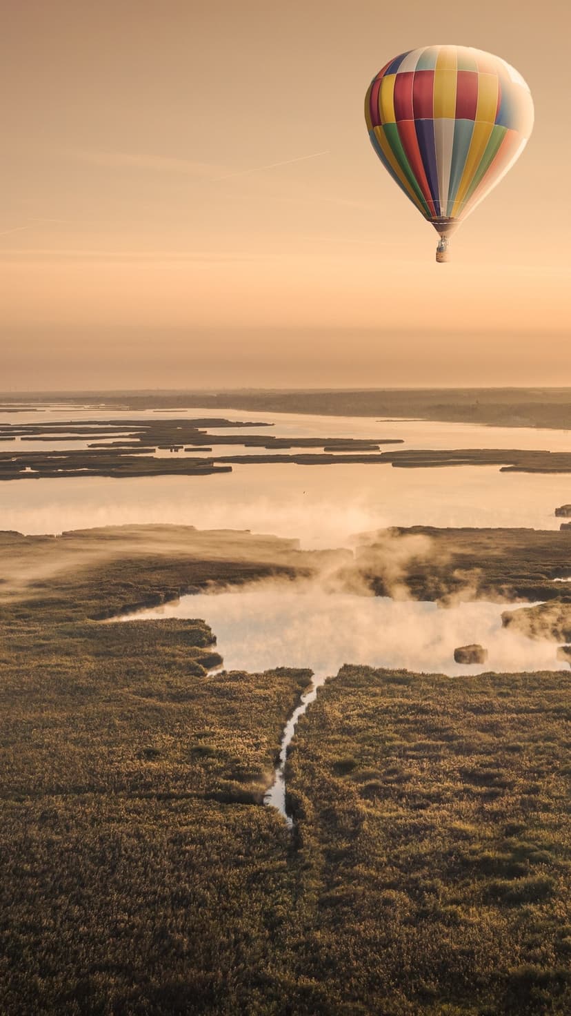 Hot air balloon soaring over winding river wetlands and rising mist under a golden sunrise in a vertical view.