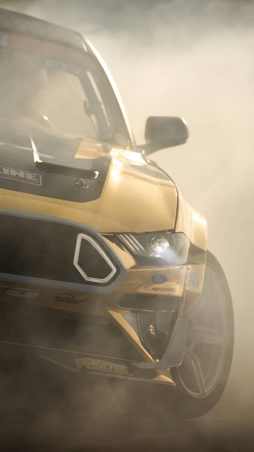 Golden Ford Mustang drifting through white smoke with racing decals visible in a low-angle vertical shot.