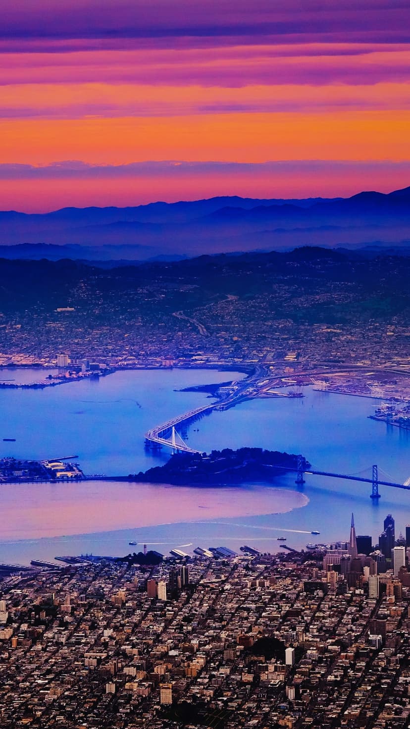 The San Francisco Bay Bridge spans blue water under an orange sky during sunset, viewed from a high altitude.