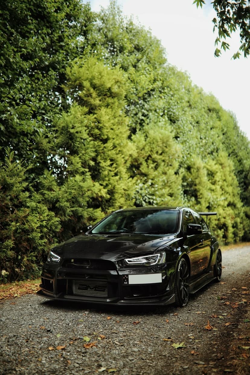 Black Mitsubishi Lancer Evolution parked on a rural gravel road surrounded by thick green forest trees.