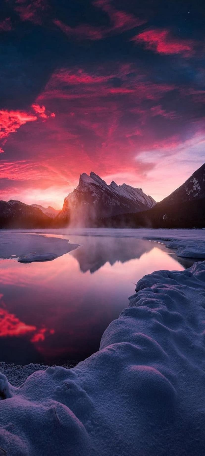 Snow-capped mountain peaks reflected in a frozen lake under a vibrant pink and purple sunset sky at twilight.