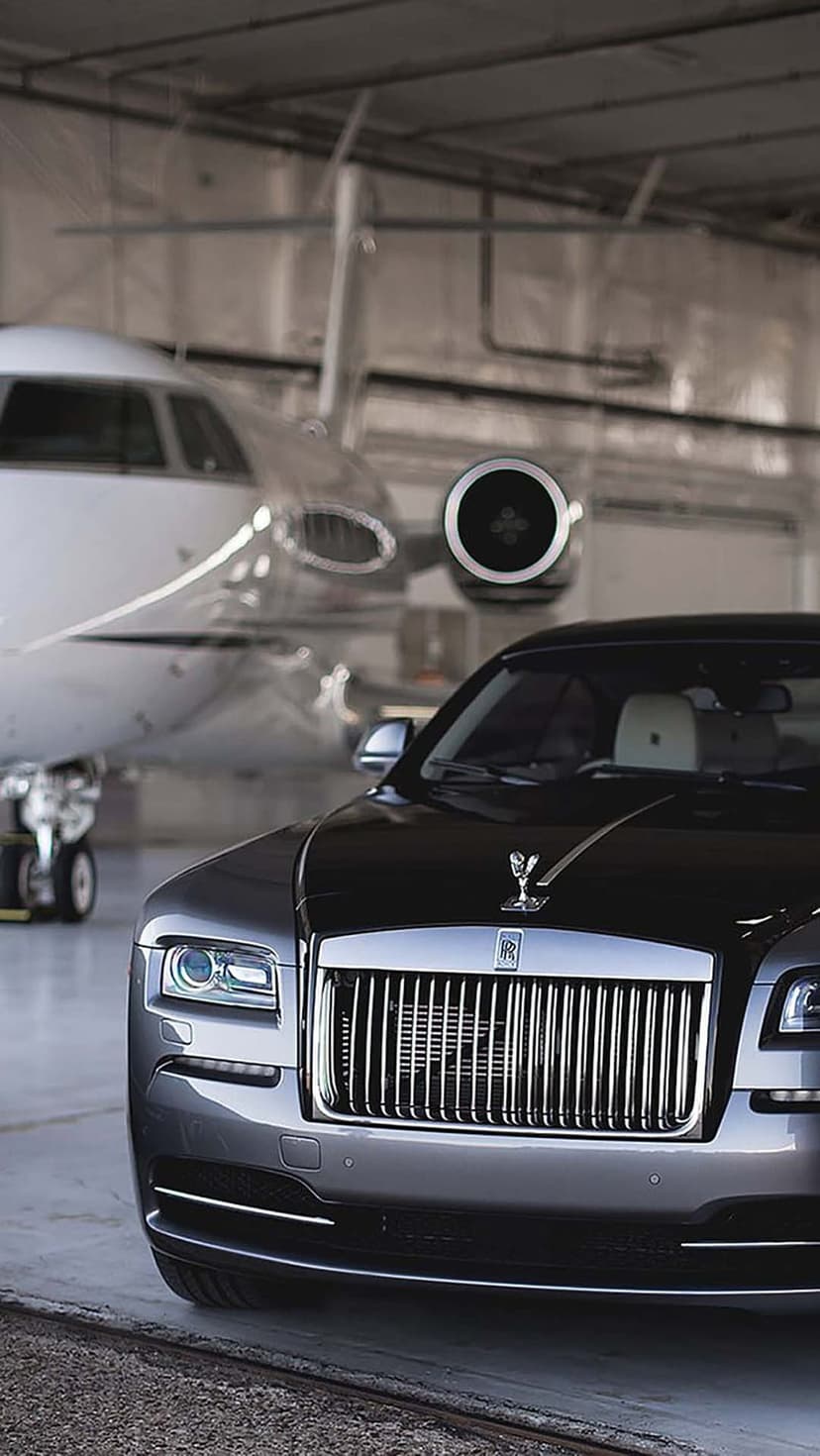 Dark grey Rolls-Royce Wraith car parked next to a private jet inside a brightly lit aircraft hangar.