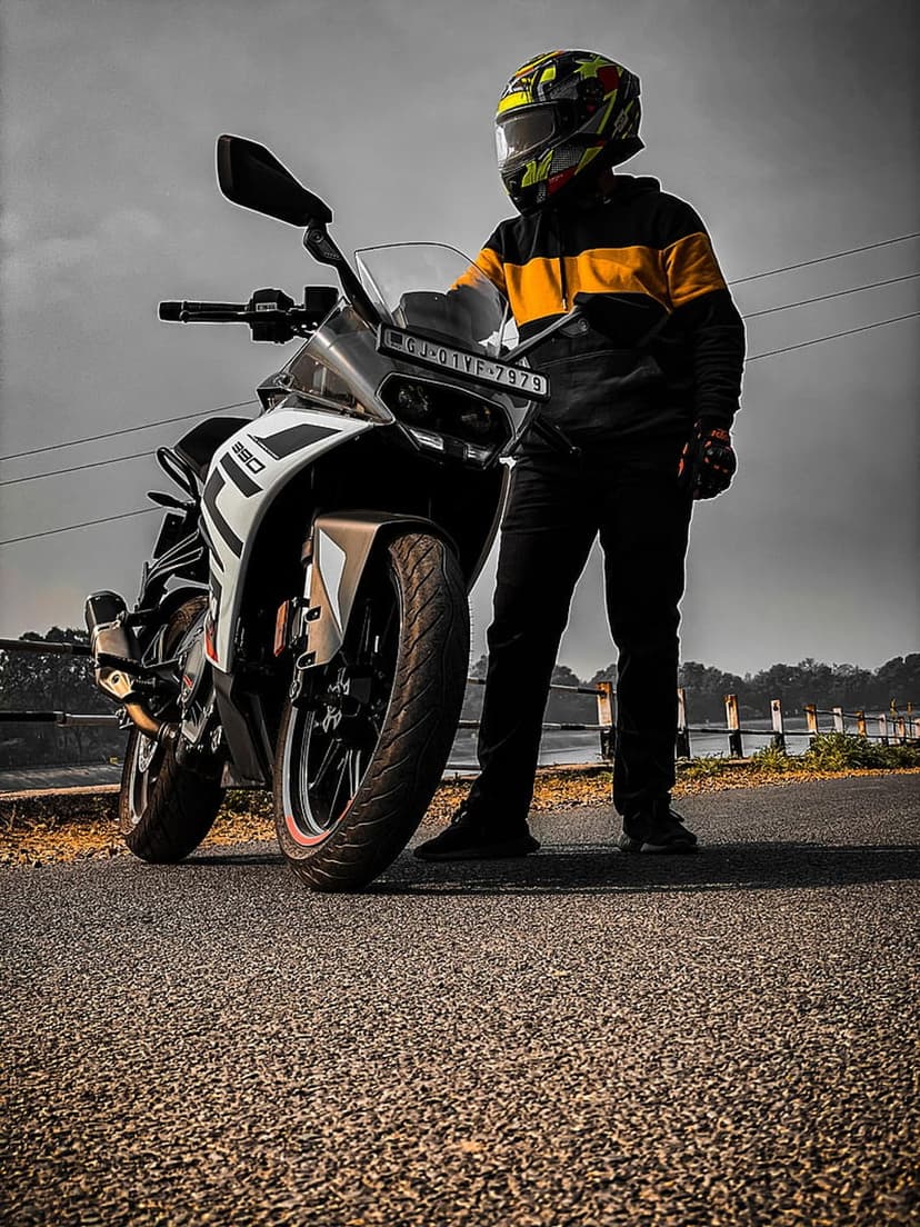 Biker in a colorful helmet stands by a white and black sportbike on a gray asphalt road under a cloudy sky.