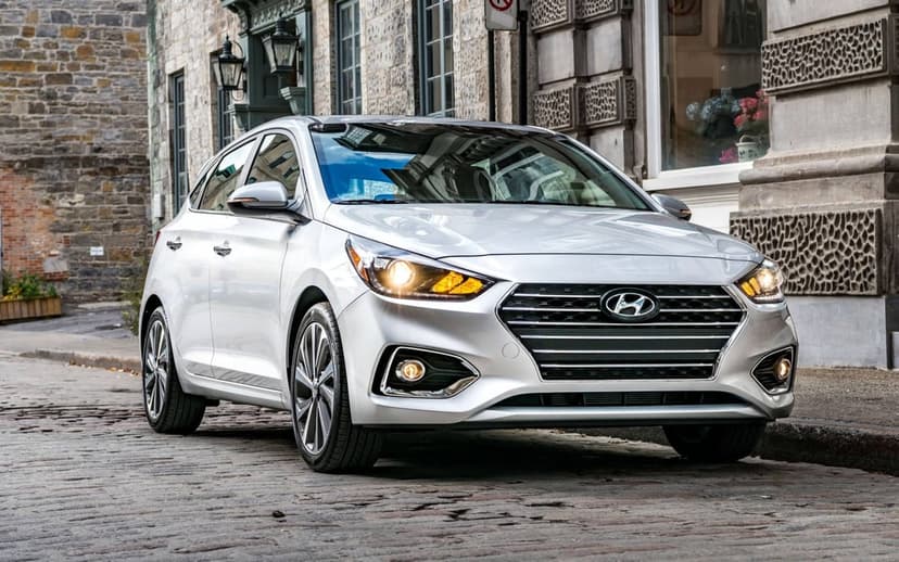Silver Hyundai Accent sedan parked on a cobblestone street with stone buildings and headlights turned on.