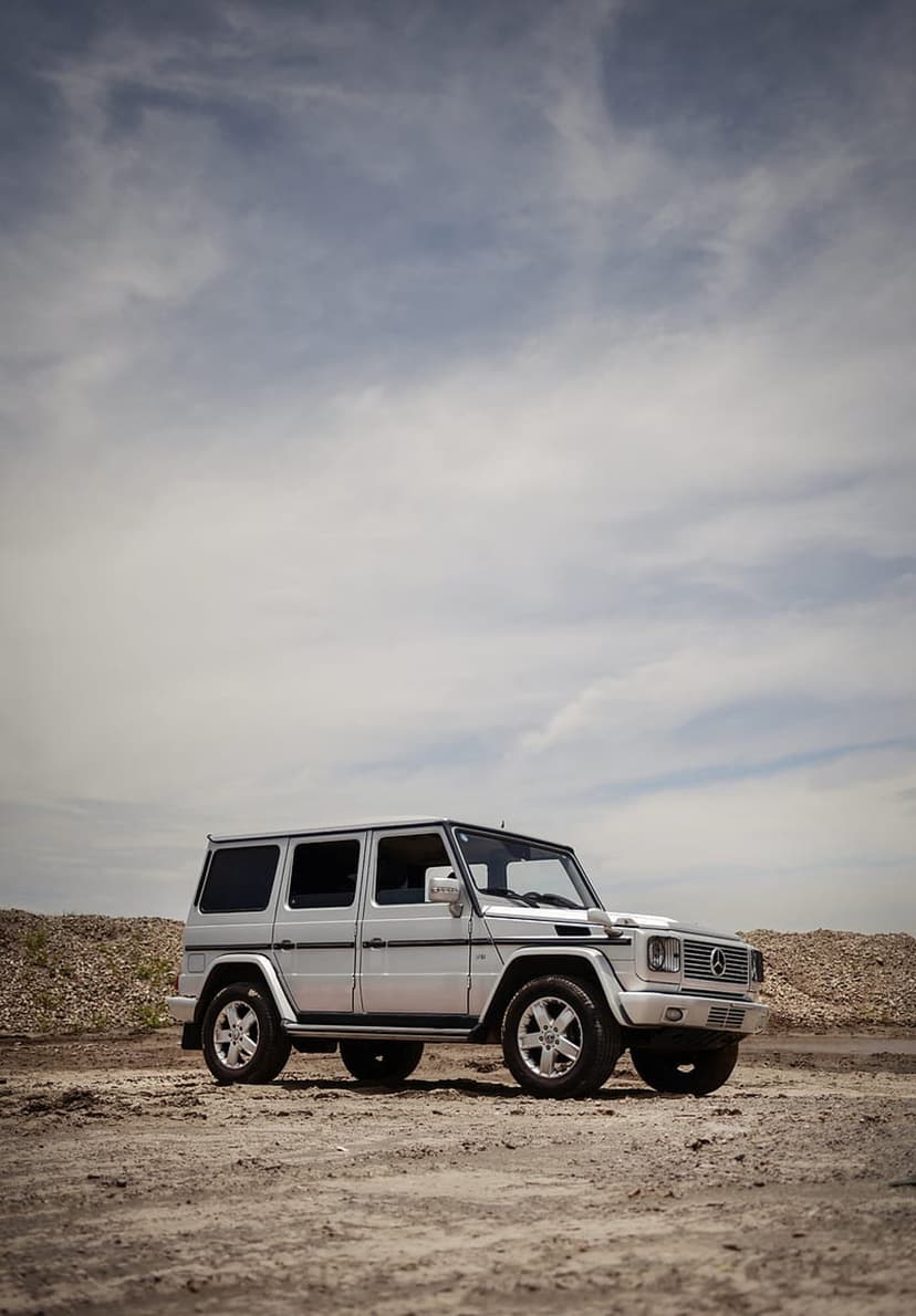 Silver Mercedes G-Class SUV parked on a gray gravel lot under an overcast sky with a boxy off-road profile.