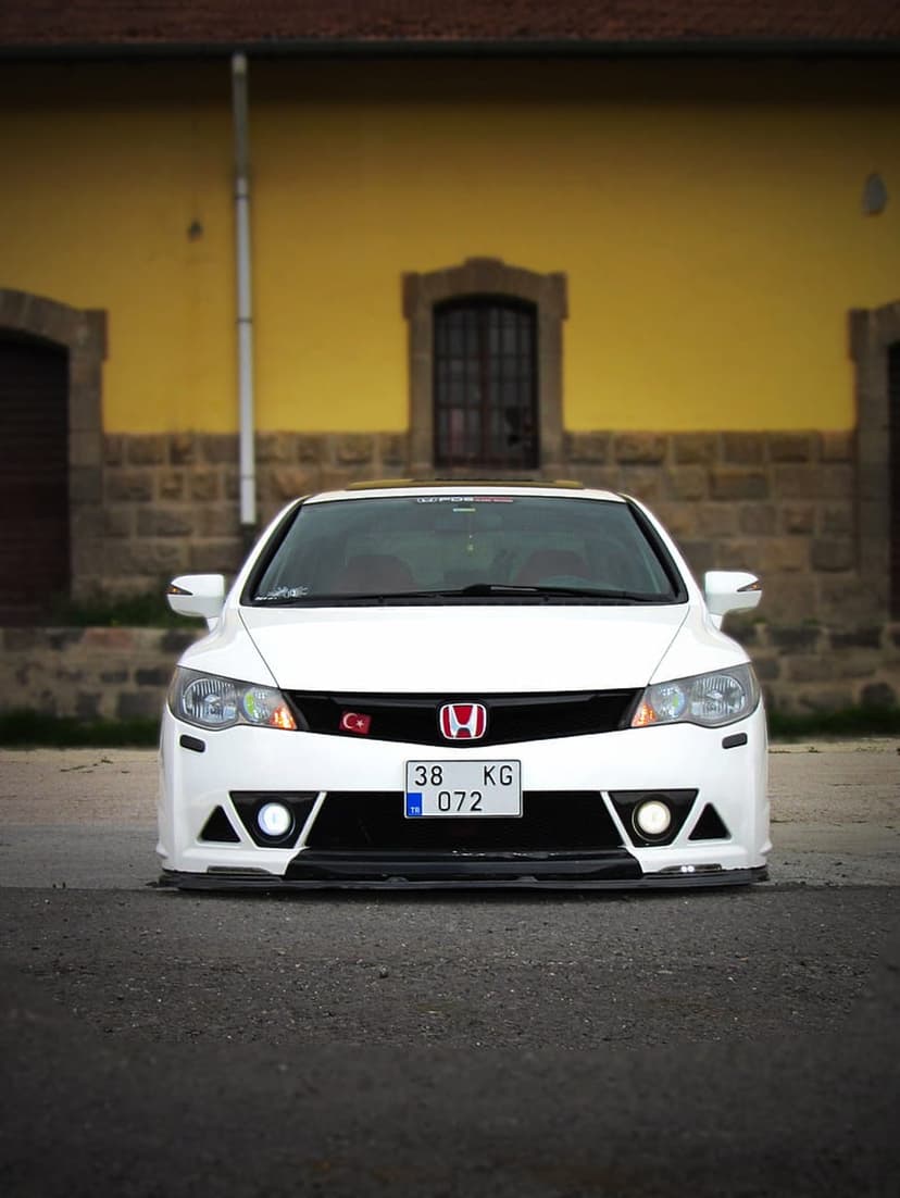 White Honda Civic with a red badge and Turkish flag parked on dark pavement in front of a yellow building.