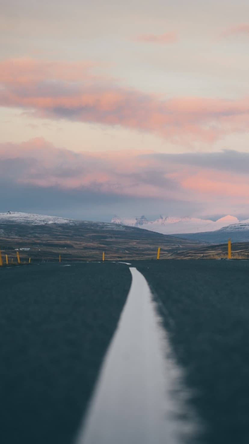 Straight asphalt road with a white center line leading toward distant snow-covered mountains under a pink sky.