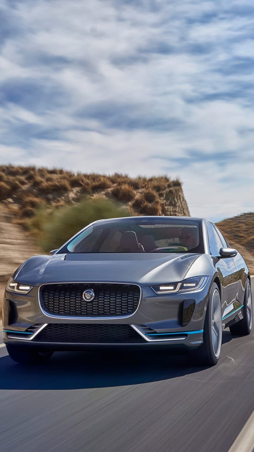 Grey Jaguar I-PACE electric SUV driving on a paved desert road with arid hills and sparse shrubs under a cloudy sky.