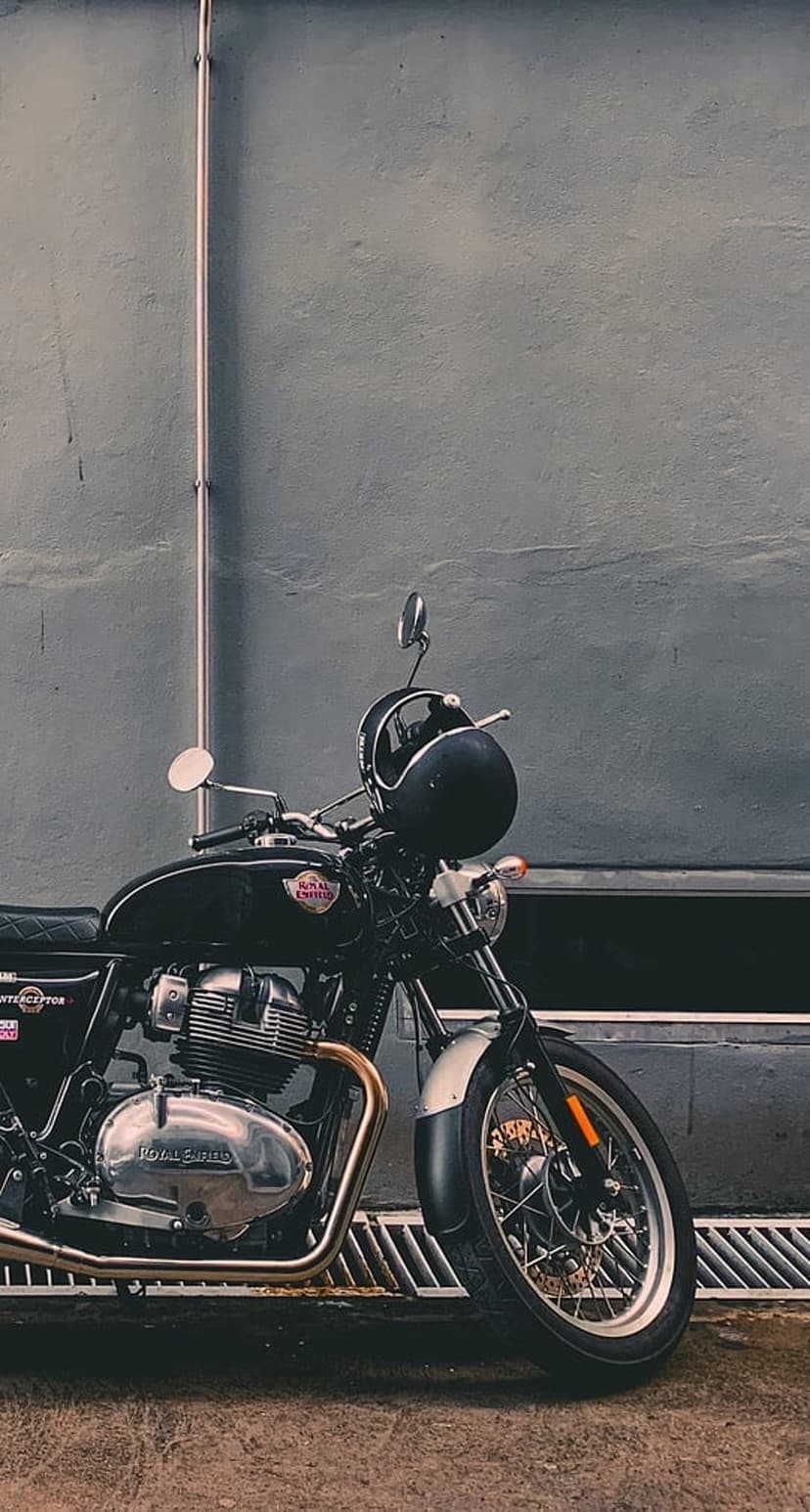 Black Royal Enfield motorcycle with a helmet on the handlebars parked against a textured grey industrial wall.