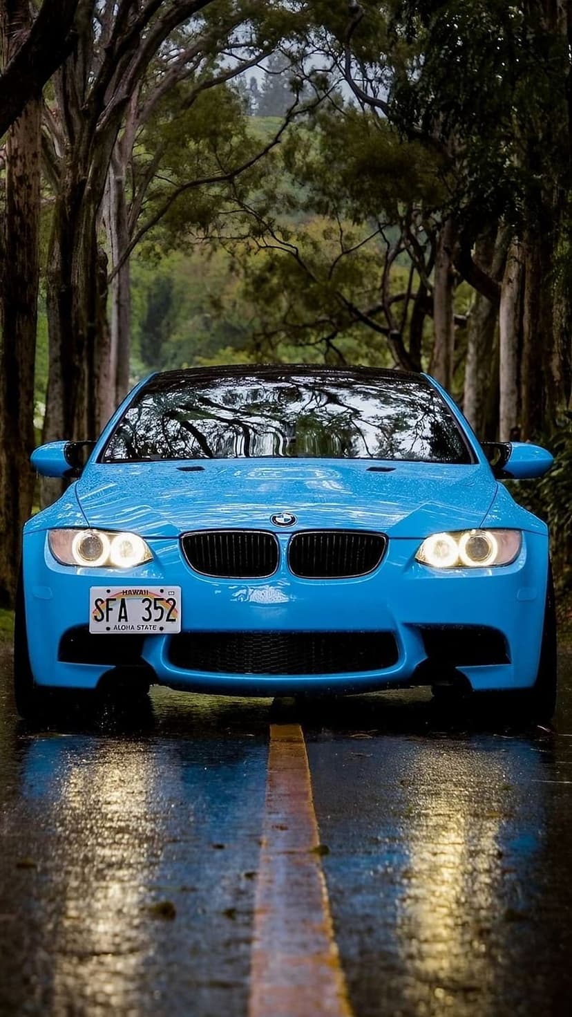 Blue BMW M3 parked on a wet asphalt road between rows of trees with its bright white headlights illuminated.