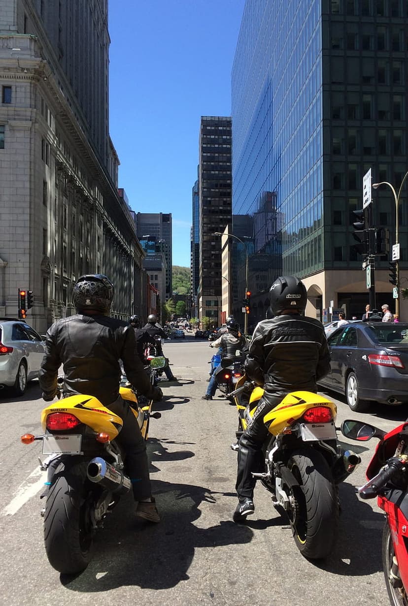 Motorcyclists in yellow gear and black leather ride sportbikes past glass skyscrapers on a sunny city street.