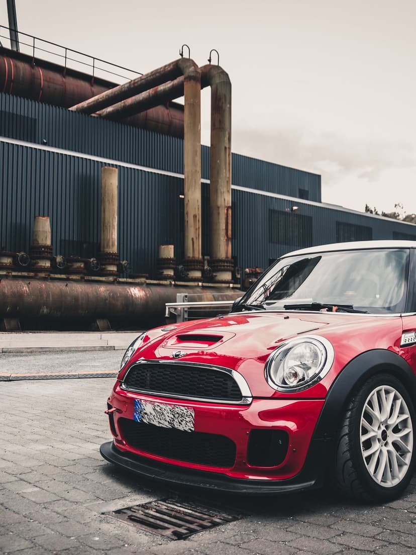 Red Mini Cooper with white wheels parked in front of large rusted industrial pipes and blue metal siding.