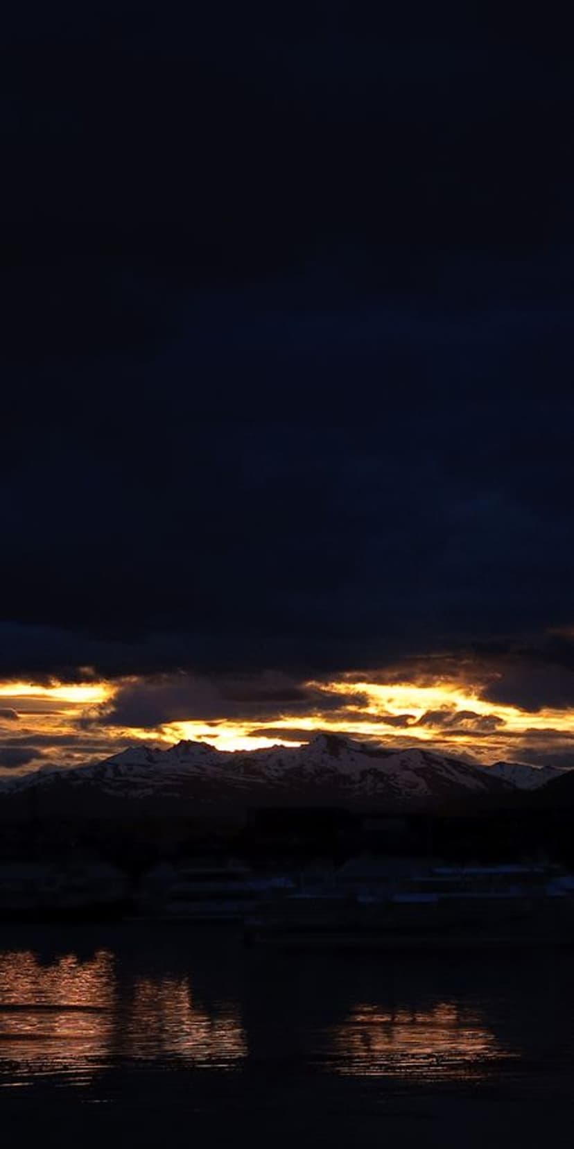 Snowy mountain peaks sit beneath a fiery orange sunset and dark storm clouds reflected in a still lake.