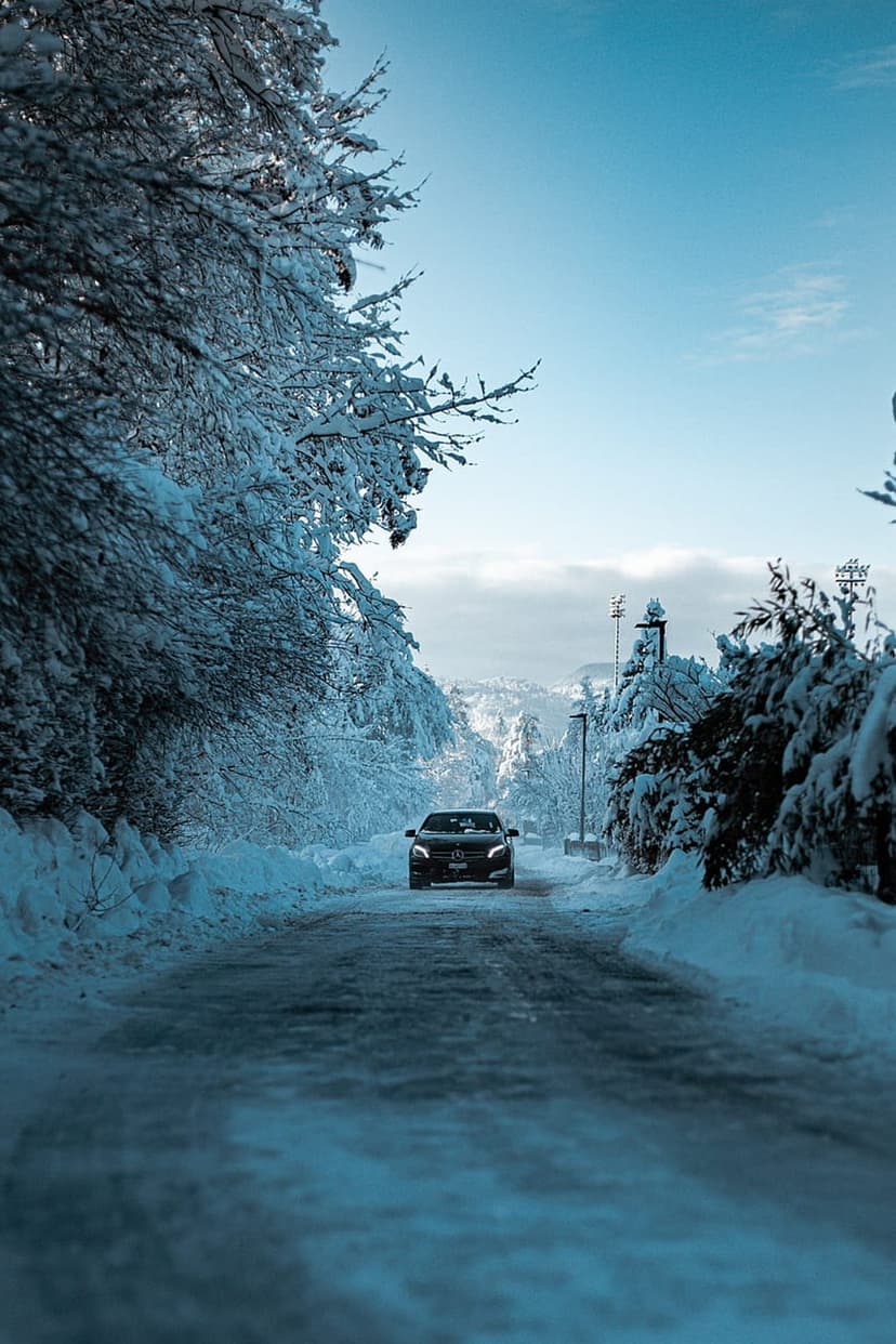 Black car drives on a snowy road flanked by frost-covered pines under a vibrant blue sky in a mountain landscape.