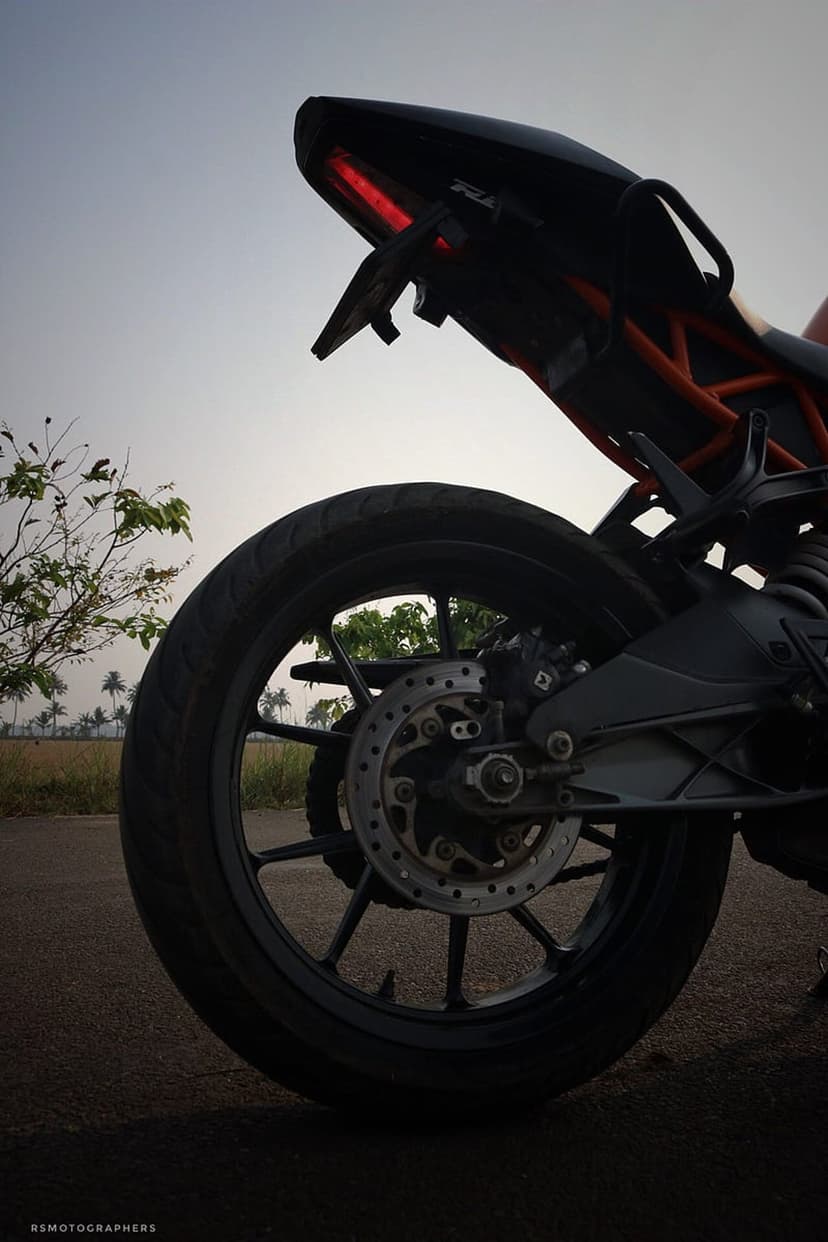 KTM motorcycle rear tire and glowing red taillight against a blurred backdrop of evening fields and trees.