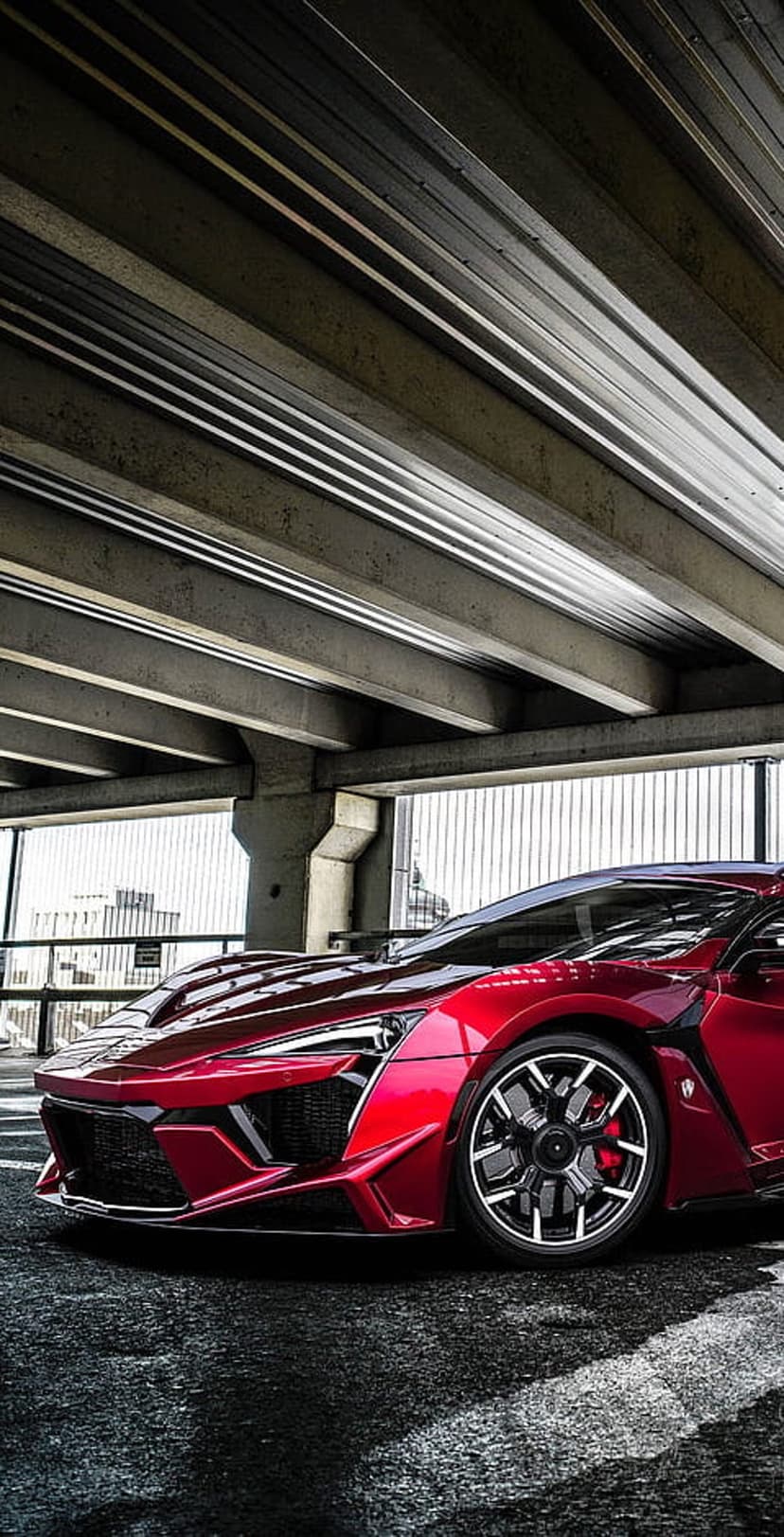 Crimson sports car parked on asphalt under a massive concrete bridge with dramatic highlights and dark shadows.