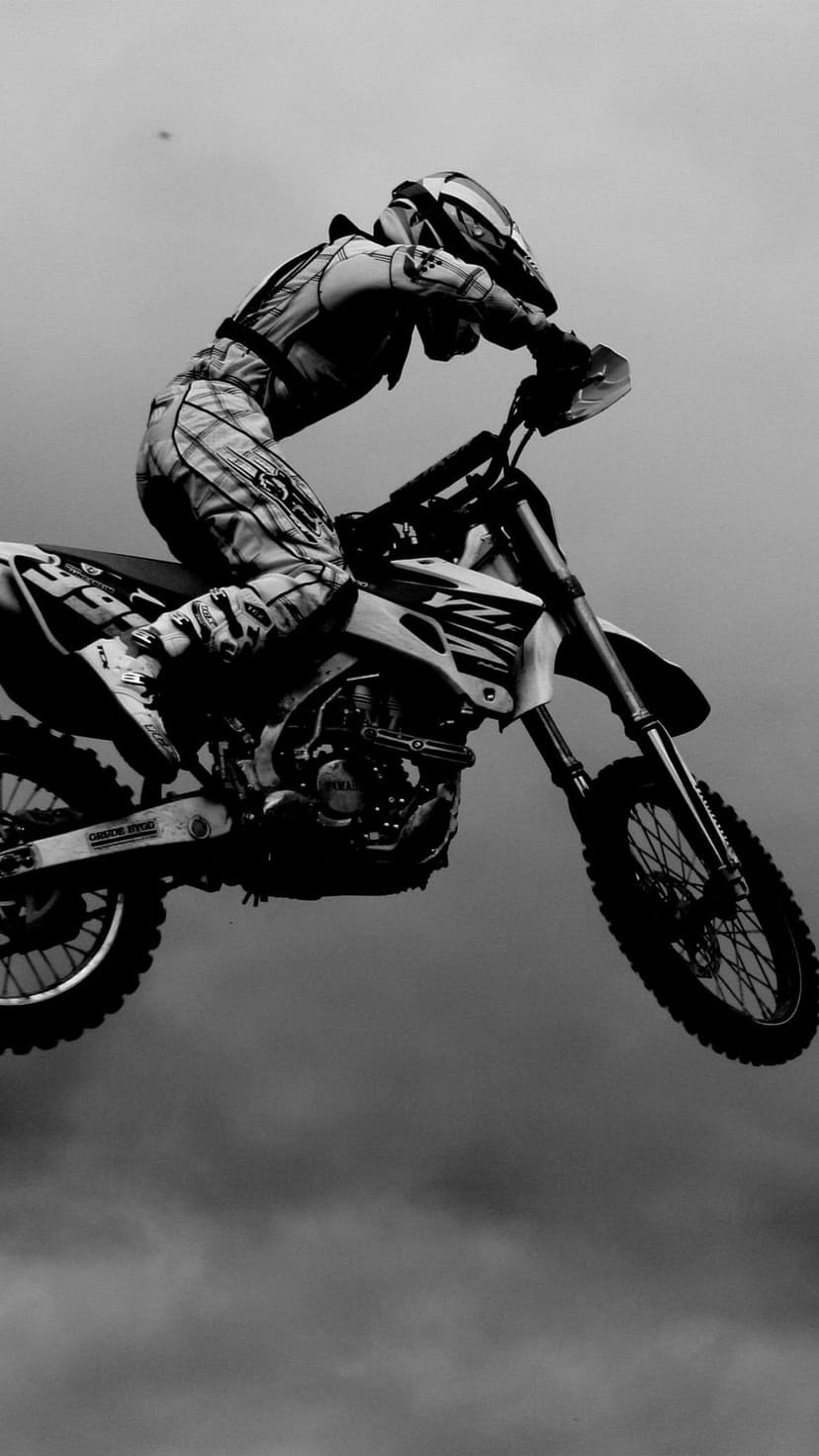 Motocross rider performing a high jump on a motorcycle against a cloudy sky in a dramatic black and white style.