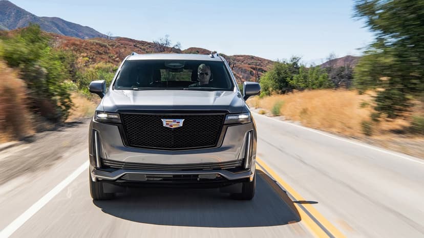 Silver Cadillac Escalade SUV driving on a paved road through golden hills under a bright blue sky with motion blur