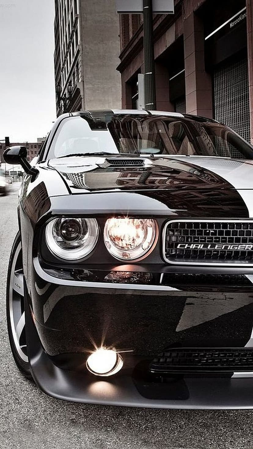Black Dodge Challenger with racing stripes parked on a brick-lined city street under a grey, cloudy sky.