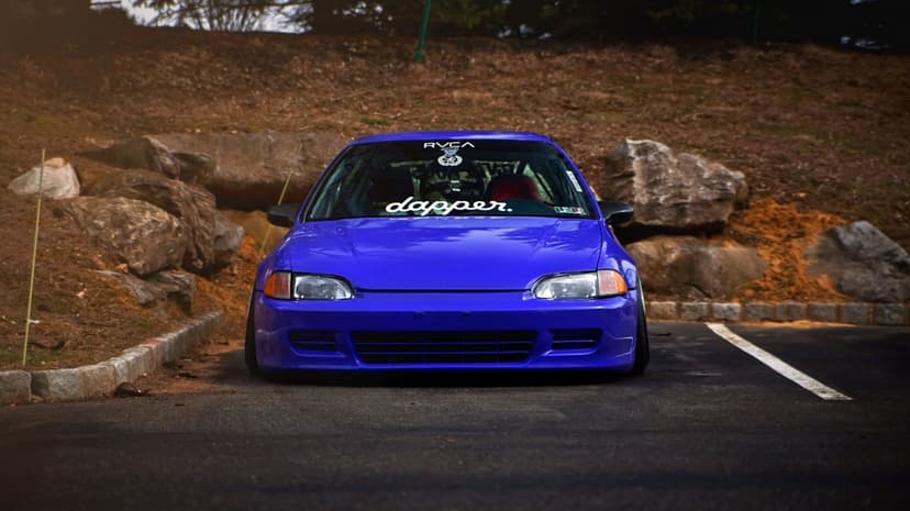 Blue Honda Civic hatchback with stanced suspension and dapper decal parked against a rocky hill background.