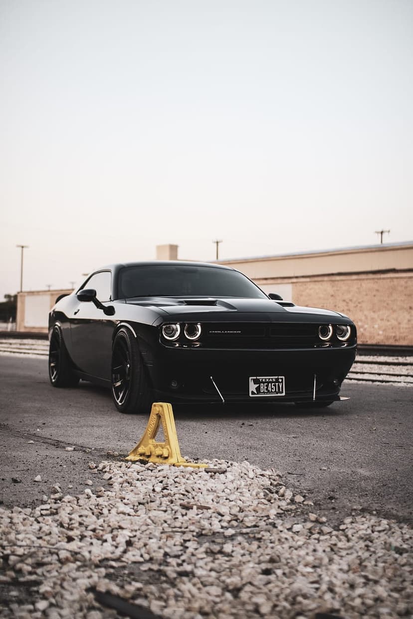 Black Dodge Challenger with a Texas BEASTY license plate parked on asphalt before an industrial backdrop.
